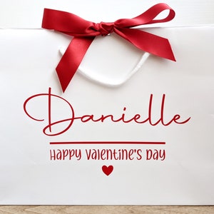 White gift bag with a red ribbon and bow. The bag has a red inscription that says "Danielle Happy Valentine's Day" with a small red heart.