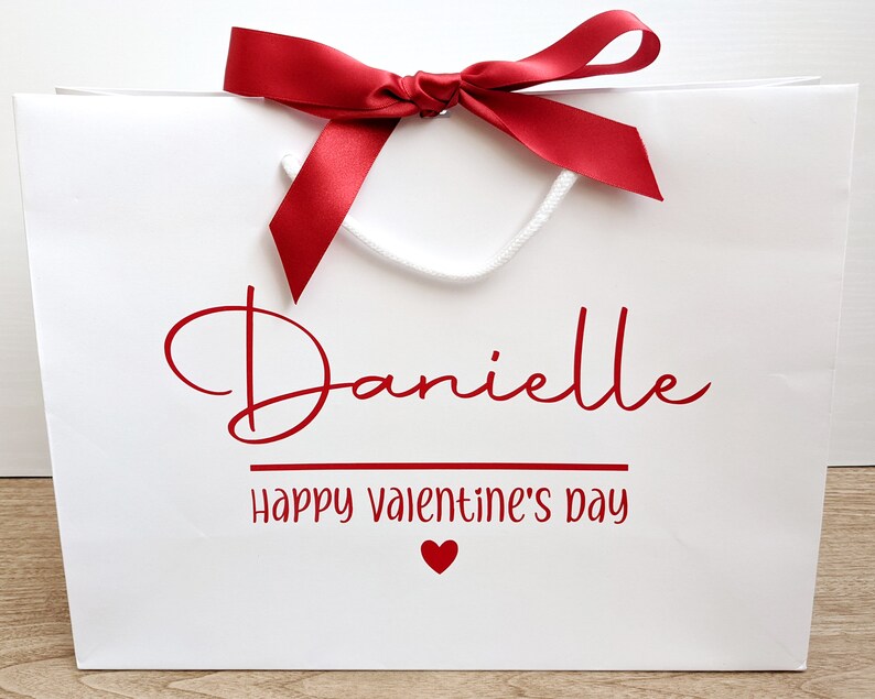 White gift bag with a red ribbon and bow. The bag has a red inscription that says "Danielle Happy Valentine's Day" with a small red heart.