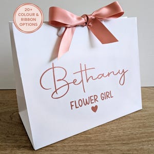 A white personalised wedding gift bag with rose gold vinyl text and rose gold satin ribbon. The name is in a large script font, with "Flower Girl" below in a smaller font.
