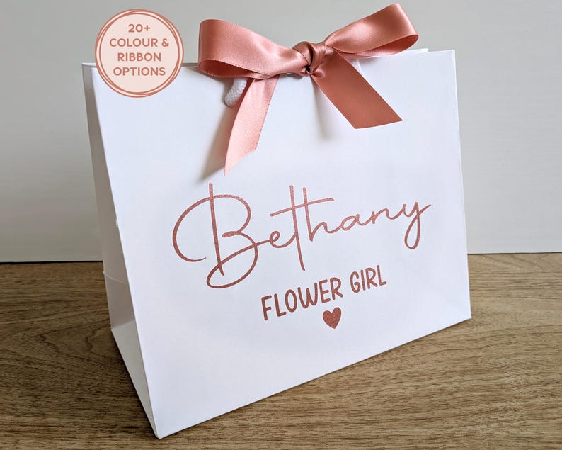 A white personalised wedding gift bag with rose gold vinyl text and rose gold satin ribbon. The name is in a large script font, with "Flower Girl" below in a smaller font.