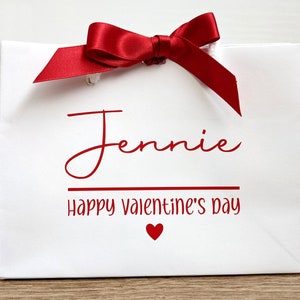 White gift bag with a red satin ribbon bow. The bag has the name "Jennie" and the text "Happy Valentine's Day" printed in red. A small red heart is printed below the text.
