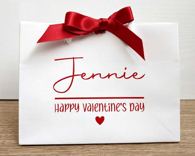 White gift bag with a red satin ribbon bow. The bag has the name "Jennie" and the text "Happy Valentine's Day" printed in red. A small red heart is printed below the text.