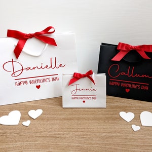 White gift bags in 3 sizes with red ribbon bows and red text. The bags are personalised with the names and the phrase "Happy Valentine's Day".
