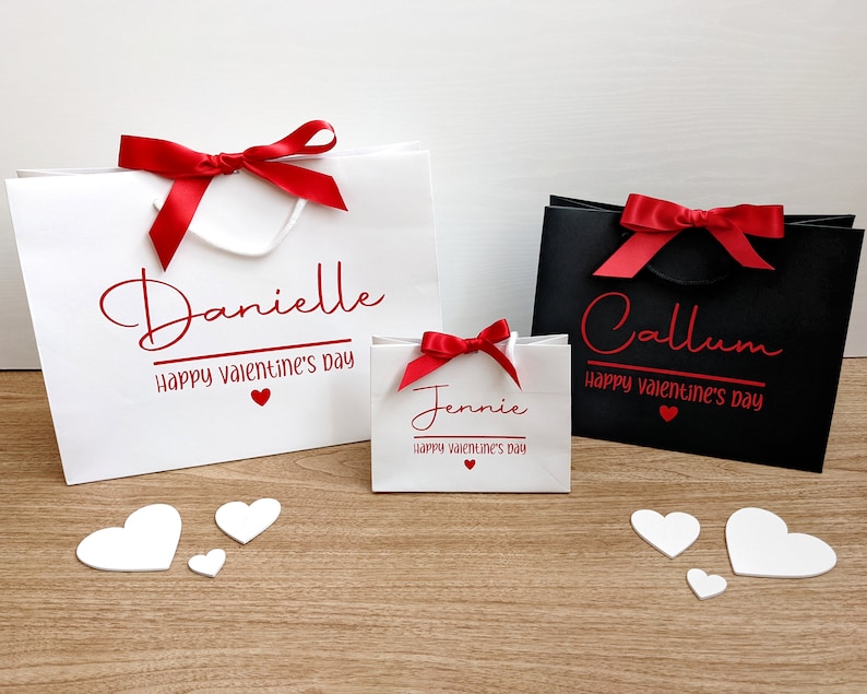 White gift bags in 3 sizes with red ribbon bows and red text. The bags are personalised with the names and the phrase "Happy Valentine's Day".
