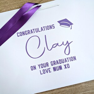 White graduation gift bag with purple text and satin ribbon.