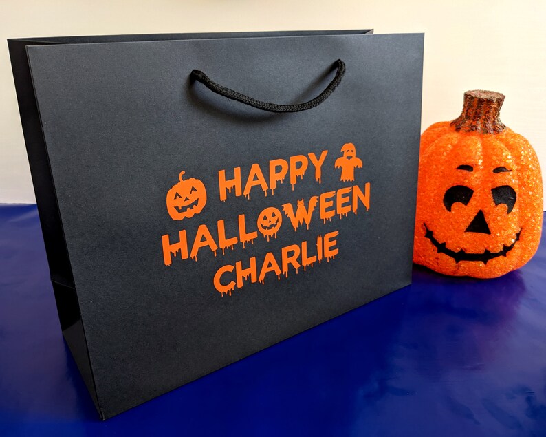 Black gift bag with orange text that says "Happy Halloween Charlie" with a pumpkin and ghost graphic.
