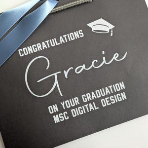 A black gift bag with blue ribbon, personalised in white text saying "Congratulations Gracie on your graduation MSc Digital Design"