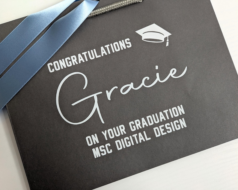 A black gift bag with blue ribbon, personalised in white text saying "Congratulations Gracie on your graduation MSc Digital Design"
