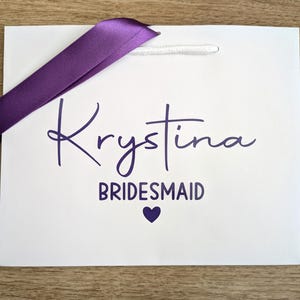 White gift bag with purple text and purple satin ribbon. The bag is personalised with a name and bridesmaid.