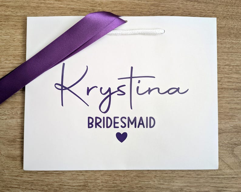 White gift bag with purple text and purple satin ribbon. The bag is personalised with a name and bridesmaid.
