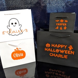 Halloween themed gift bags with personalised names. The first bag is white with a ghost and the name in orange. The second bag is white with a pumpkin and the name in orange. The third bag is black with orange text that says Happy Halloween and a name and includes a pumpkin, ghost, and bat graphic.