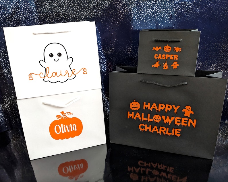 Halloween themed gift bags with personalised names. The first bag is white with a ghost and the name in orange. The second bag is white with a pumpkin and the name in orange. The third bag is black with orange text that says Happy Halloween and a name and includes a pumpkin, ghost, and bat graphic.