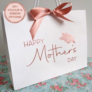 White gift bag reading Happy Mother's Day with a floral motif at the top right of the design. Bag shown has text in rose gold and rose gold ribbon.