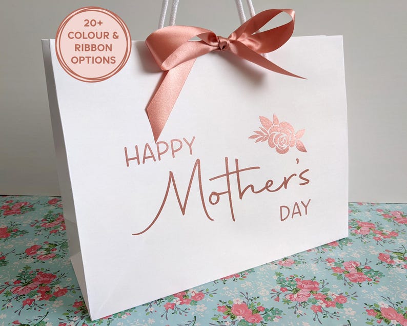 White gift bag reading Happy Mother's Day with a floral motif at the top right of the design. Bag shown has text in rose gold and rose gold ribbon.