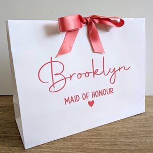 A white personalised gift bag with a name in large text and Maid of Honour below in smaller text. The text and ribbon are a matching coral colour.