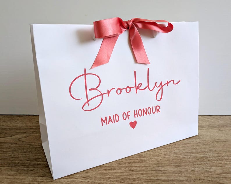 A white personalised gift bag with a name in large text and Maid of Honour below in smaller text. The text and ribbon are a matching coral colour.