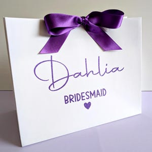 A white gift bag with purple glitter vinyl text and ribbon. The name is in a large cursive font with "Bridesmaid" below in smaller text.