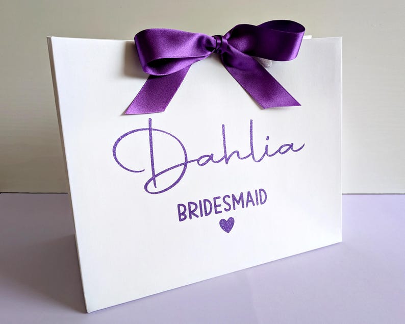 A white gift bag with purple glitter vinyl text and ribbon. The name is in a large cursive font with "Bridesmaid" below in smaller text.