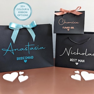 3 black gift bags in different sizes. Personalised with a name in decorative font, with wedding role below in smaller font. One bag has blue text, one has rose gold and one has white. Two of the bags also have ribbon.