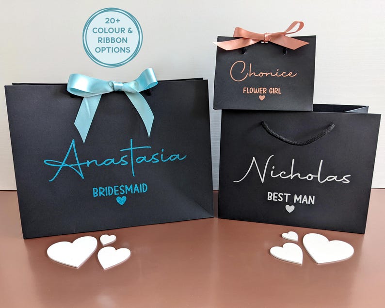 3 black gift bags in different sizes. Personalised with a name in decorative font, with wedding role below in smaller font. One bag has blue text, one has rose gold and one has white. Two of the bags also have ribbon.