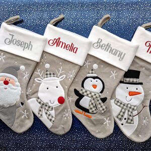 May include: Four Christmas stockings, each with a name embroidered on the cuff: Joseph, Amelia, Bethany, and Theo. The stockings feature Santa, a reindeer, a penguin, and a snowman. Beige stockings with white cuffs and festive designs.