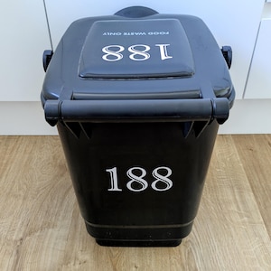 May include: A black plastic waste bin featuring a hinged lid and a handle. The number 188 is printed in white on both the front of the bin and the top of the lid. The text FOOD WASTE ONLY is printed in white on the top lid above the number 188.