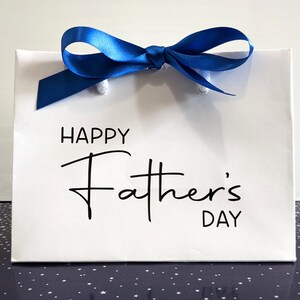 White gift bag with a blue ribbon and the text "Happy Father's Day" on the front in black.