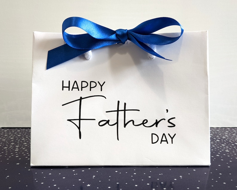 White gift bag with a blue ribbon and the text "Happy Father's Day" on the front in black.