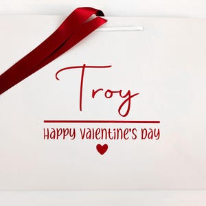 A white gift bag with red writing and ribbon. It reads "Troy Happy Valentine's Day".