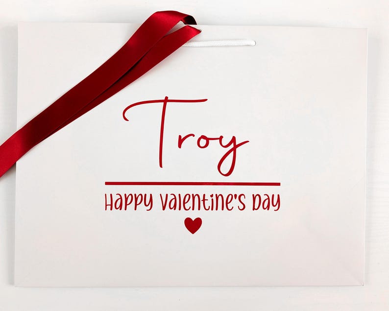 A white gift bag with red writing and ribbon. It reads "Troy Happy Valentine's Day".