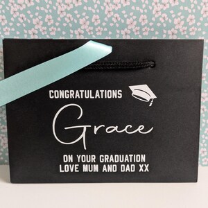 A black gift bag with turquoise ribbon, personalised in white text which says "Congratulations Grace on your graduation love Mum and Dad xx"