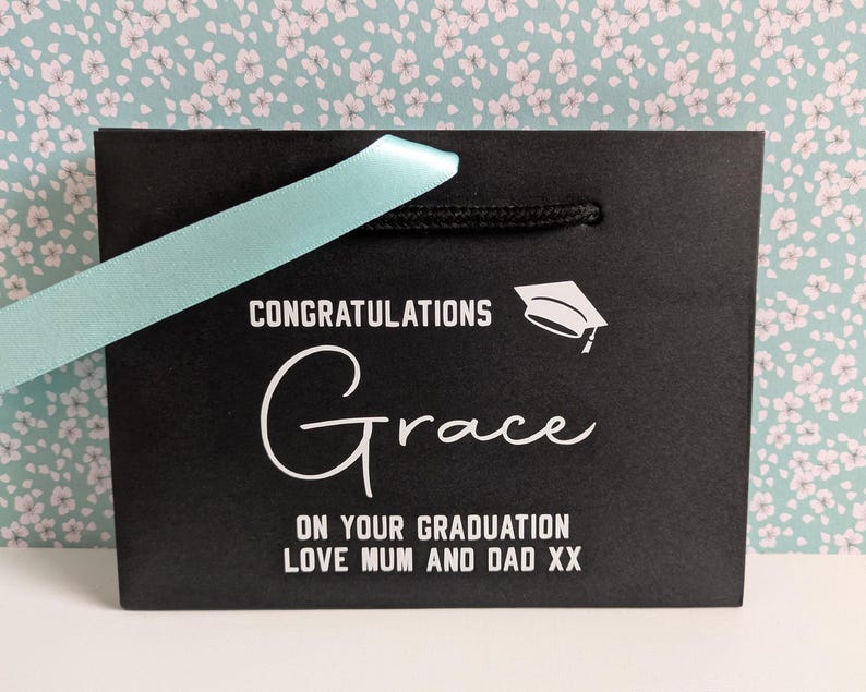 A black gift bag with turquoise ribbon, personalised in white text which says "Congratulations Grace on your graduation love Mum and Dad xx"