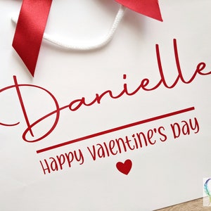 White gift bag with a red ribbon and the text "Danielle Happy Valentine's Day" in red cursive font. A small red heart is below the text.