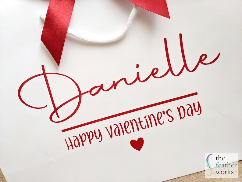 White gift bag with a red ribbon and the text "Danielle Happy Valentine's Day" in red cursive font. A small red heart is below the text.