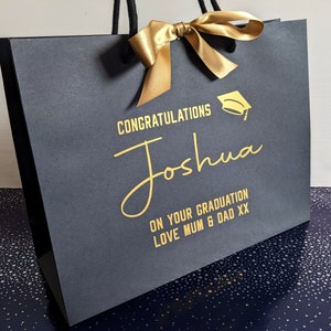 Black gift bag with a gold ribbon and a gold graduation cap graphic. The bag has the text "Congratulations Joshua On Your Graduation Love Mum & Dad XX" printed on it.