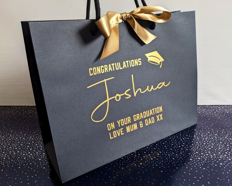 Black gift bag with a gold ribbon and a gold graduation cap graphic. The bag has the text "Congratulations Joshua On Your Graduation Love Mum & Dad XX" printed on it.