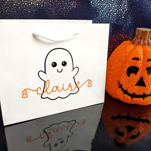 White gift bag with a black and white ghost illustration and the name written in orange. The bag has a white handle and is sitting on a blue background with a small orange pumpkin.
