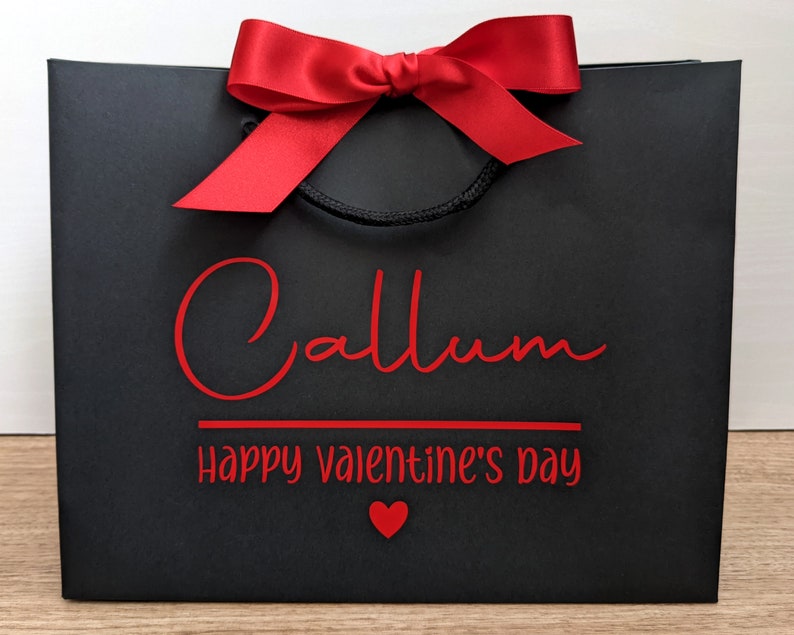 Black gift bag with a red bow and the text "Callum Happy Valentine's Day" in red lettering.