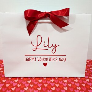 A white gift bag with red ribbon bow and red text. It has the name "Lily" in red text, with the message "Happy Valentine's Day" below.