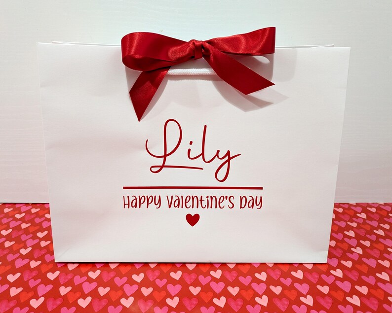 A white gift bag with red ribbon bow and red text. It has the name "Lily" in red text, with the message "Happy Valentine's Day" below.