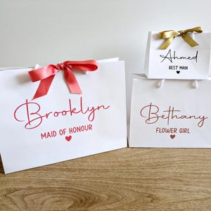 3 white gift bags with a wedding theme. They all have a name in large text, with the wedding role below in smaller text. The text is in a range of colours, these bags have coral, rose gold and black text, and coral and gold satin ribbons.
