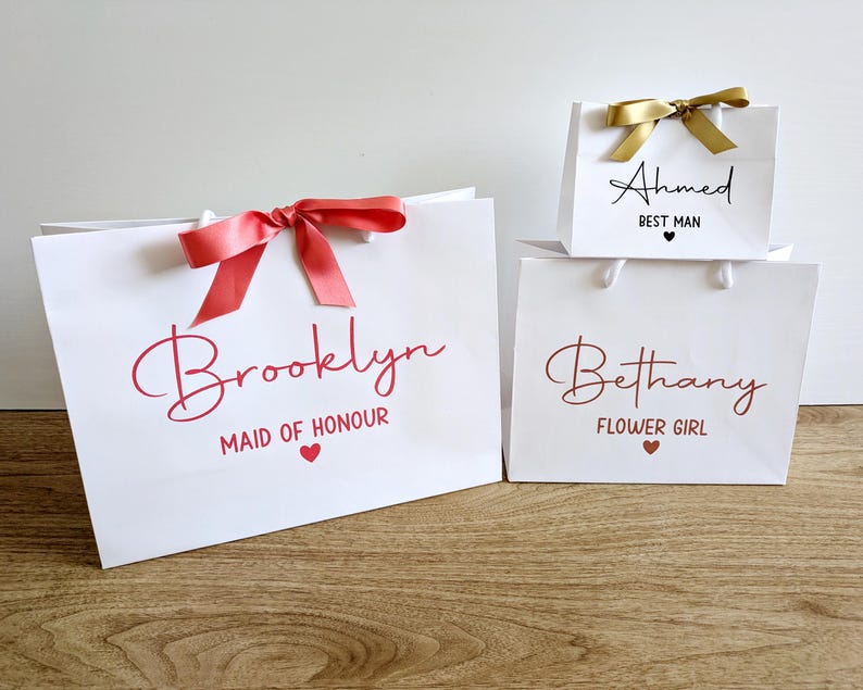 3 white gift bags with a wedding theme. They all have a name in large text, with the wedding role below in smaller text. The text is in a range of colours, these bags have coral, rose gold and black text, and coral and gold satin ribbons.