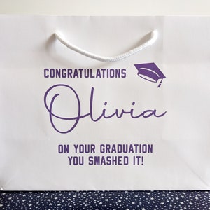 White gift bag with a purple graduation cap and the text "Congratulations Olivia on your graduation you smashed it!"
