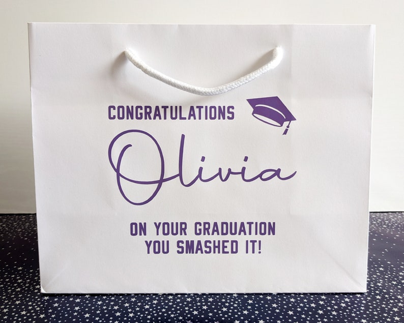 White gift bag with a purple graduation cap and the text "Congratulations Olivia on your graduation you smashed it!"