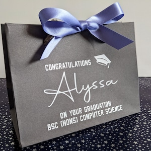 A grey gift bag with a blue ribbon and a white graduation cap graphic. The bag is personalised in white vinyl and says "Congratulations Alyssa on your graduation, BSc Hons Computer Science"