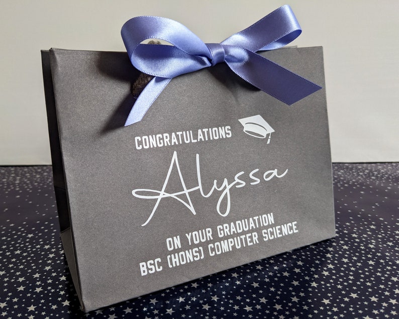 A grey gift bag with a blue ribbon and a white graduation cap graphic. The bag is personalised in white vinyl and says "Congratulations Alyssa on your graduation, BSc Hons Computer Science"