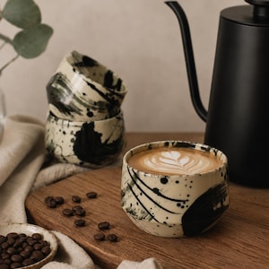 Könnte beinhalten: Drei Keramiktassen mit cremefarbener Basis und schwarzen Farbklecksen sowie Pinselstrichmustern. Eine Tasse ist mit einem Latte gefüllt, der weißen Milchschaum zeigt. Ein schwarzer Schwanenhals-Wasserkocher und geröstete Kaffeebohnen sind zu sehen.