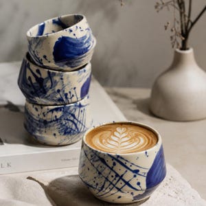 Op de afbeelding: Een set van vier keramische bekers met een crèmekleurige basis en handgeschilderde blauwe abstracte penseelstreken en spetters. Eén beker is gevuld met een latte met een bladvormig schuimpatroon, terwijl de andere drie verticaal zijn gestapeld.