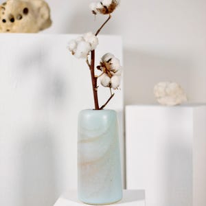 Könnte beinhalten: Eine zylindrische Keramikvase mit einer glatten, hellblauen Glasur und dezenten, wirbelnden beigen und weißen Marmormustern. Ein brauner Stiel mit mehreren flauschigen, weißen Baumwollkapseln steckt in der Vase und ragt nach oben.