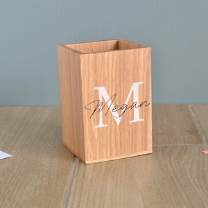Könnte beinhalten: Ein hellbrauner Schreibtisch-Organizer aus Holz. Er hat einen großen weißen Buchstaben "M" und den Namen "Megan" in Schreibschrift. Der Artikel ist quadratisch und steht auf einer Holzoberfläche.